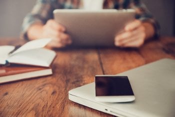 Laptop and cell phone on a table with a person holding a tablet in the background.