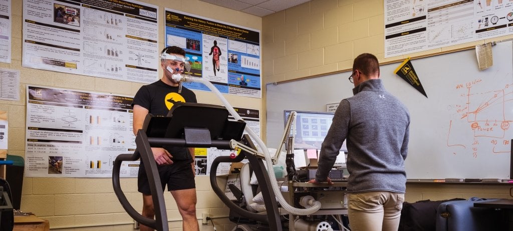 Researcher connects a blood pressure cuff to a student standing on a treadmill.