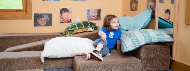 Young child sitting on pillows in a classroom.