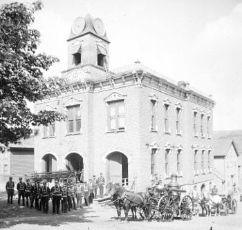 Historical photo of the fire hall with the fire fighters and horses outside.