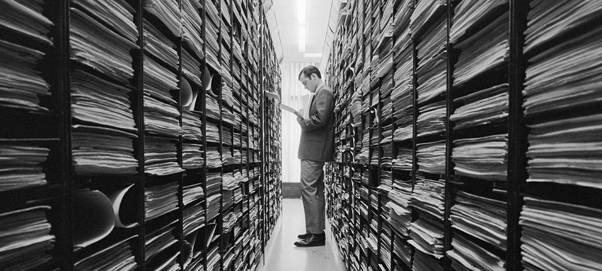 Person standing between shelves of files looking at papers.