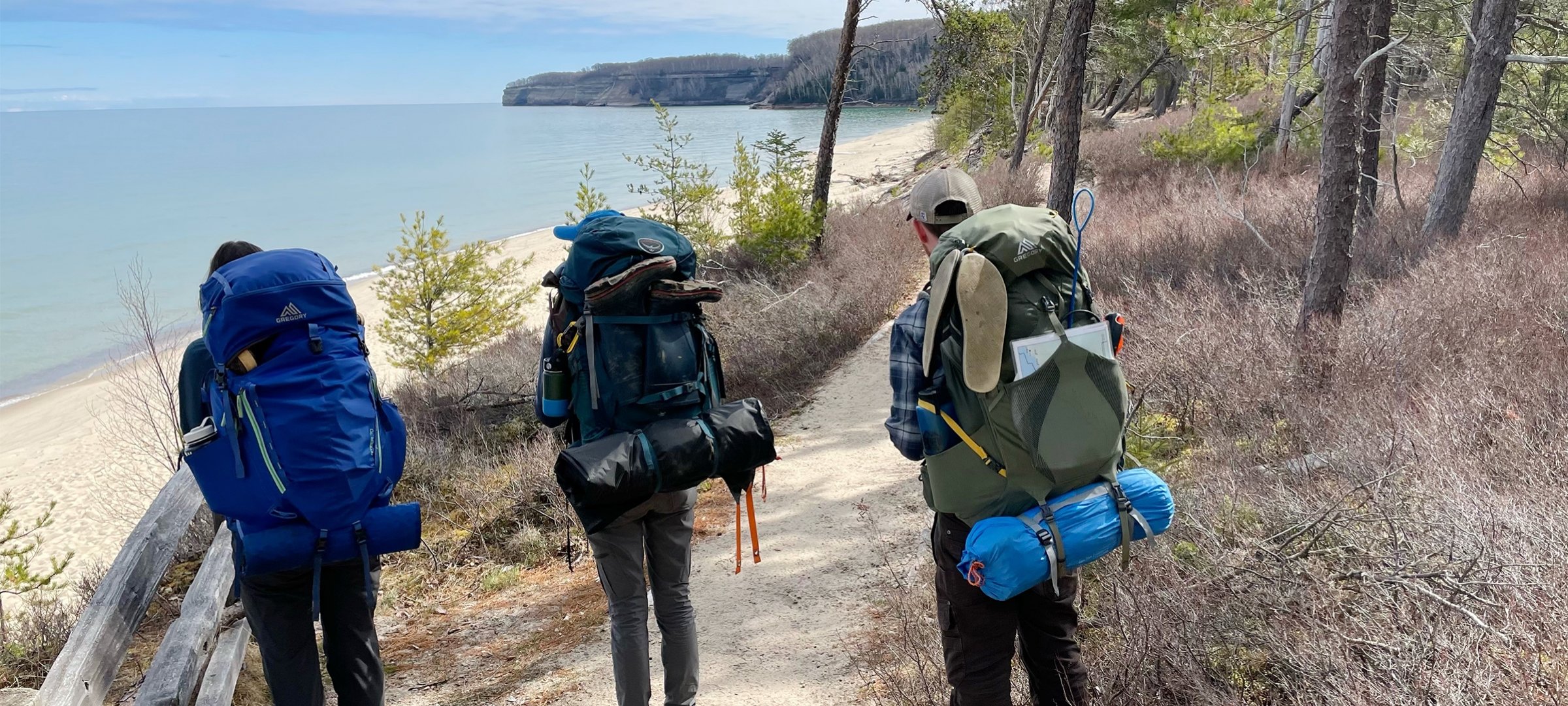 Three people with hiking backpacks looking out over a beach and water.