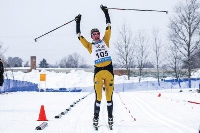 Deedra Irwin, wearing a Michigan Tech uniform, lifts her arms in celebration at the end of a ski race.