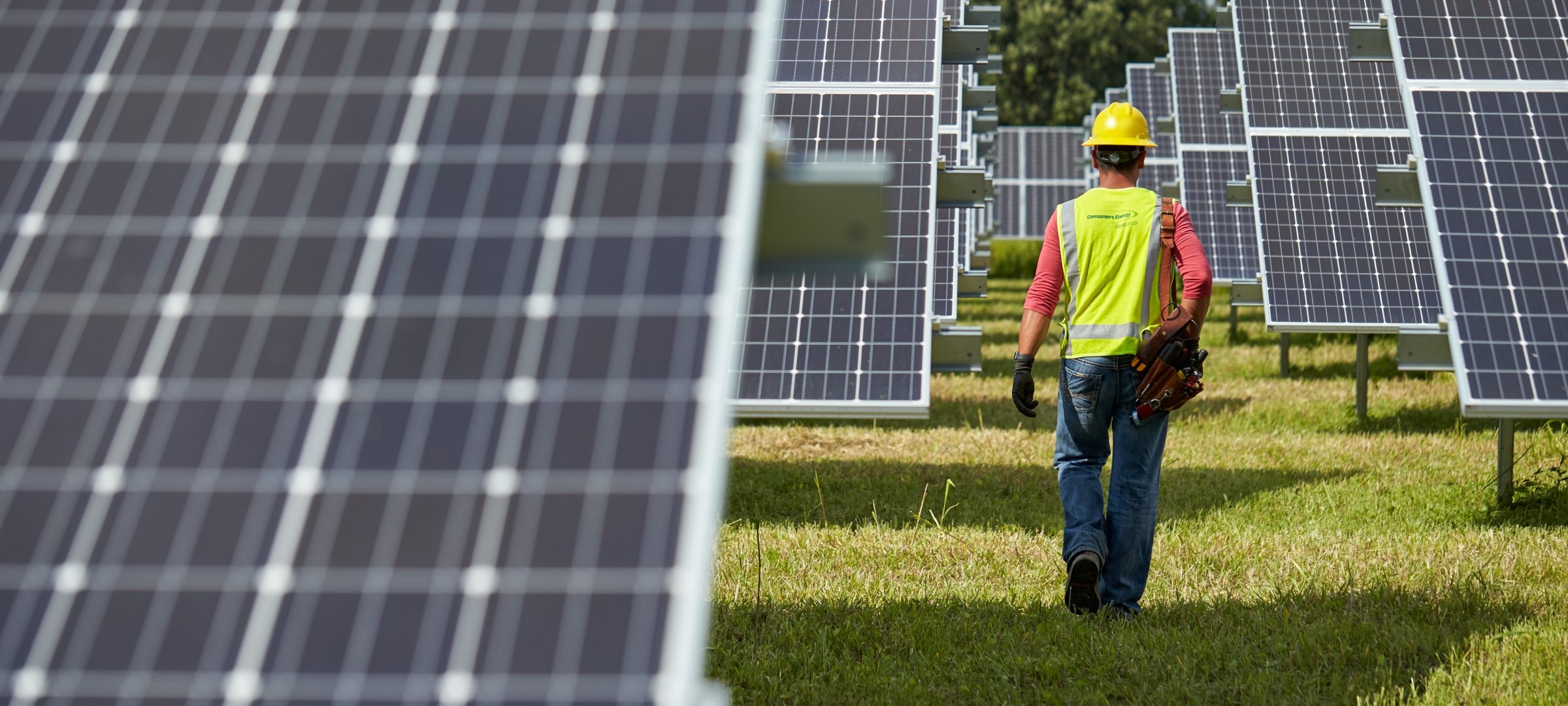 Person in a hardhat and Consumers Energy vest walking between rows of solar panels.