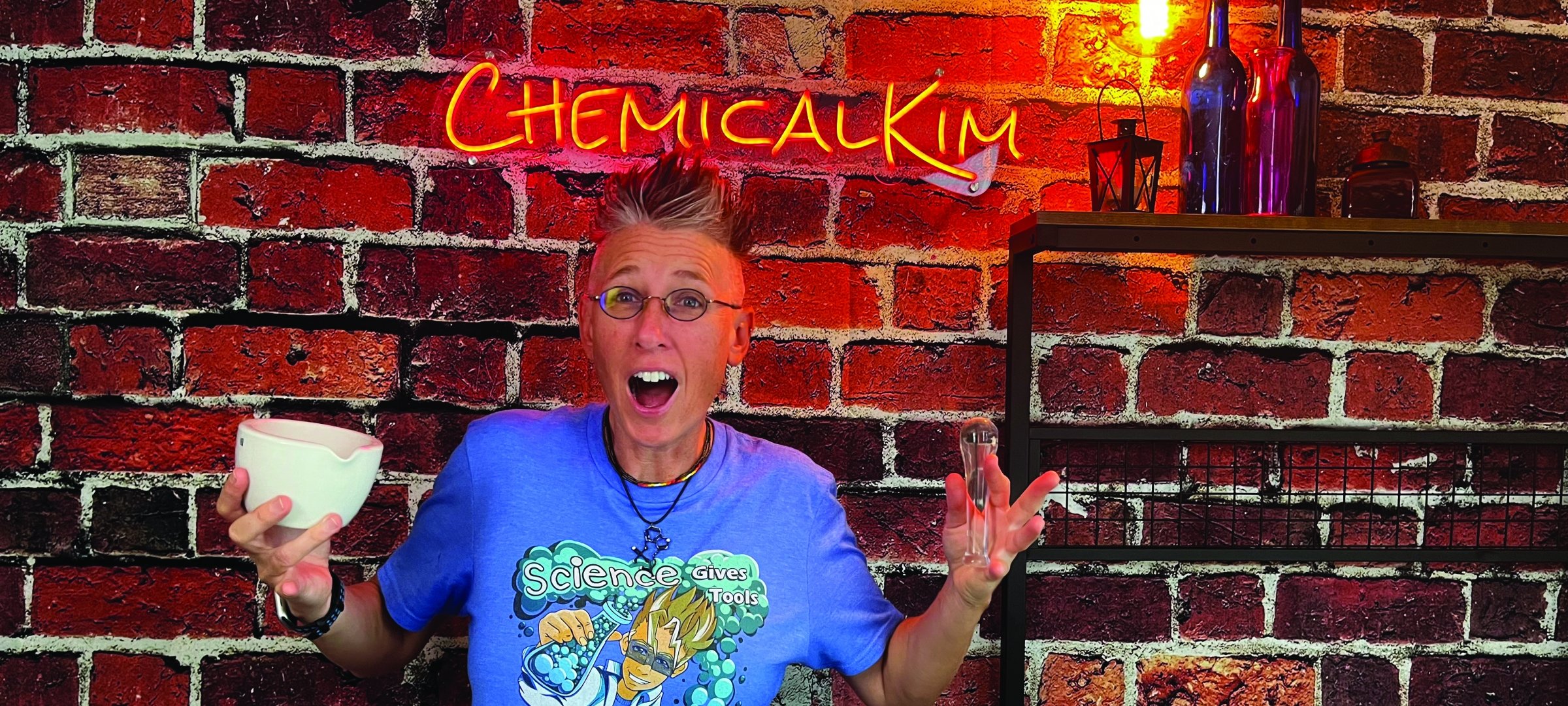 Chemical Kim standing in front of a brick wall with neon Chemical Kim sign holding a bowl and test tube.