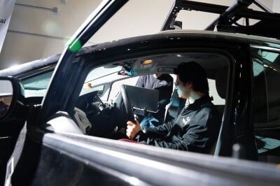 Researcher sitting in the driver's seat of a test car while another has a laptop standing outside the passenger side.