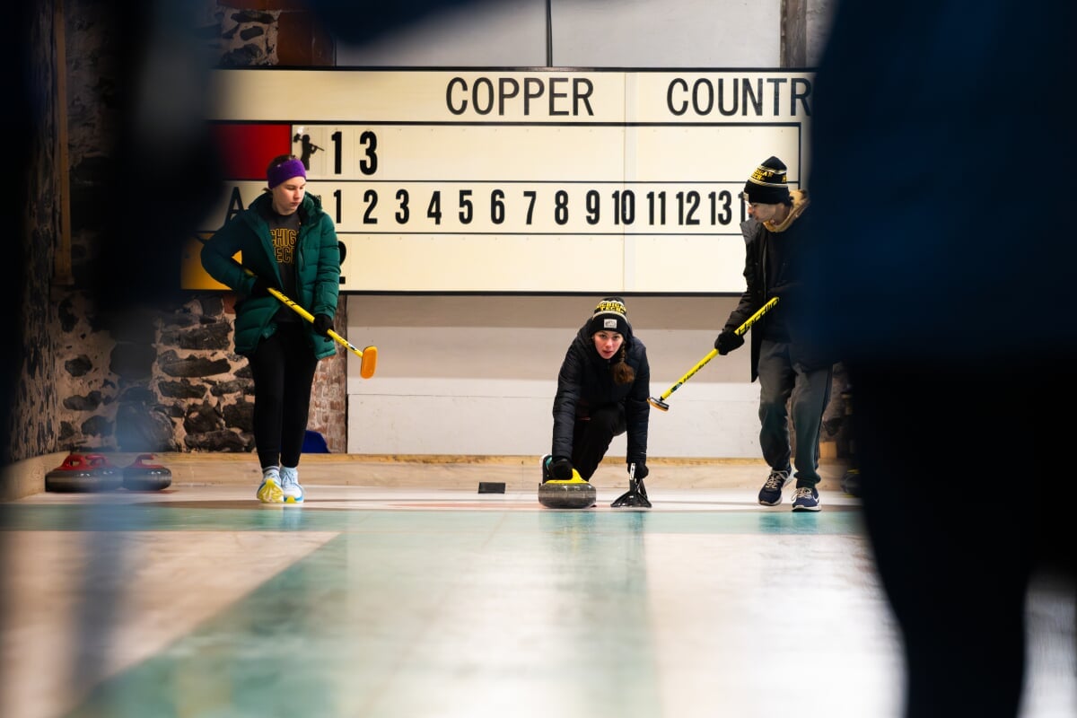 Student releasing a curling stone during a game while two others stand ready with brooms.