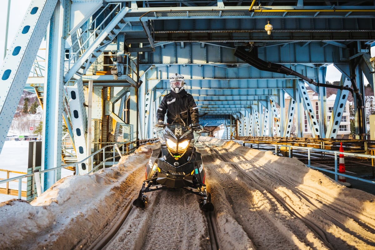 A snowmobile driving toward the camera on the trail under the Portage Lake Lift Bridge.