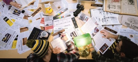 Papers, camera, water bottles clutter a table while students work around it.