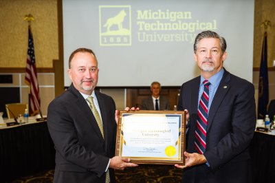 Two men holding a framed award between them with the Michigan Tech logo on a screen in the background.
