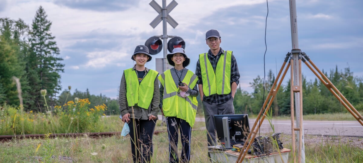 Three students in safety vests stand at a railroad crossing.