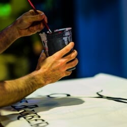 Hnads holding a cup of paint over a white sheet.