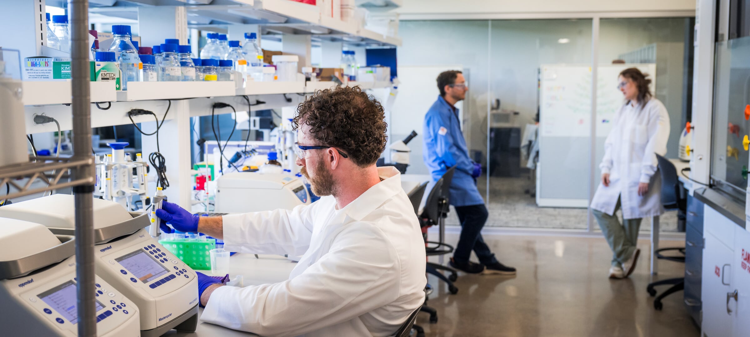 Researcher processing samples in a lab while two others converse in the background.