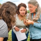 A group of women gather around a teacher who holds up a small plant.