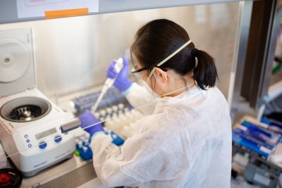 A person wearing full protective gear uses a pipette in a lab hood.