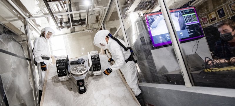 Group of people in protective gear stand next to a lunar rover inside a vinyl and polycarbonate-walled chamber.