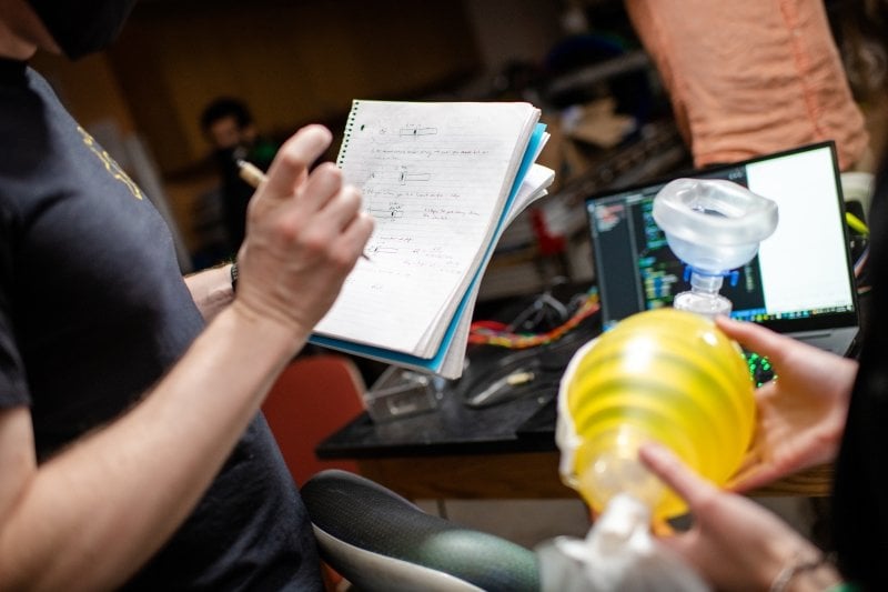 Two researchers, one writing, one holding the 3D-printed ventilator