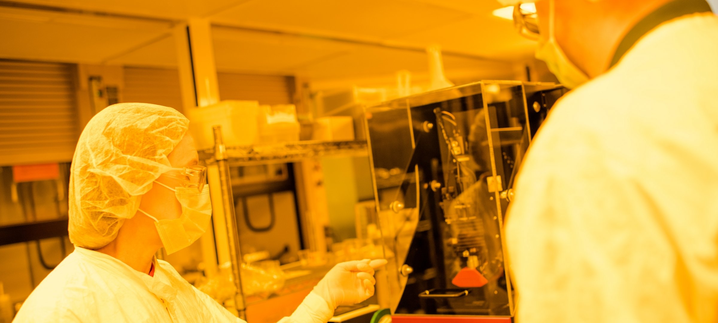 Two researchers in goggles, masks, head and clothes coverings inside Microfabrication Lab pointing to a piece of equipment