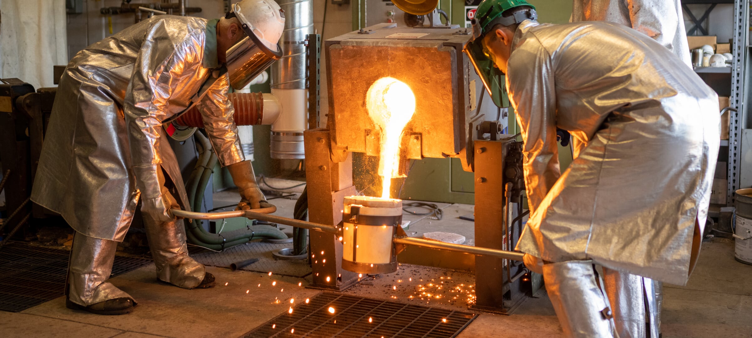Two researcher wearing full heat protective gear pour liquid metal into a container.