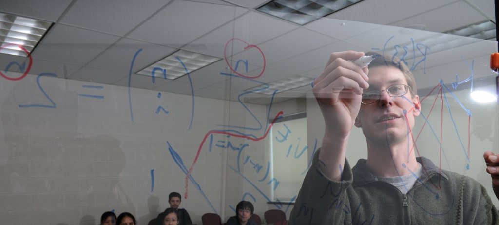 Student instructing seated students while writing equations on a clear board.