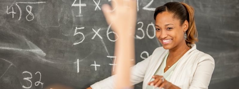 Elementary School Teacher standing in front of a chalkboard selecting raised hands of students