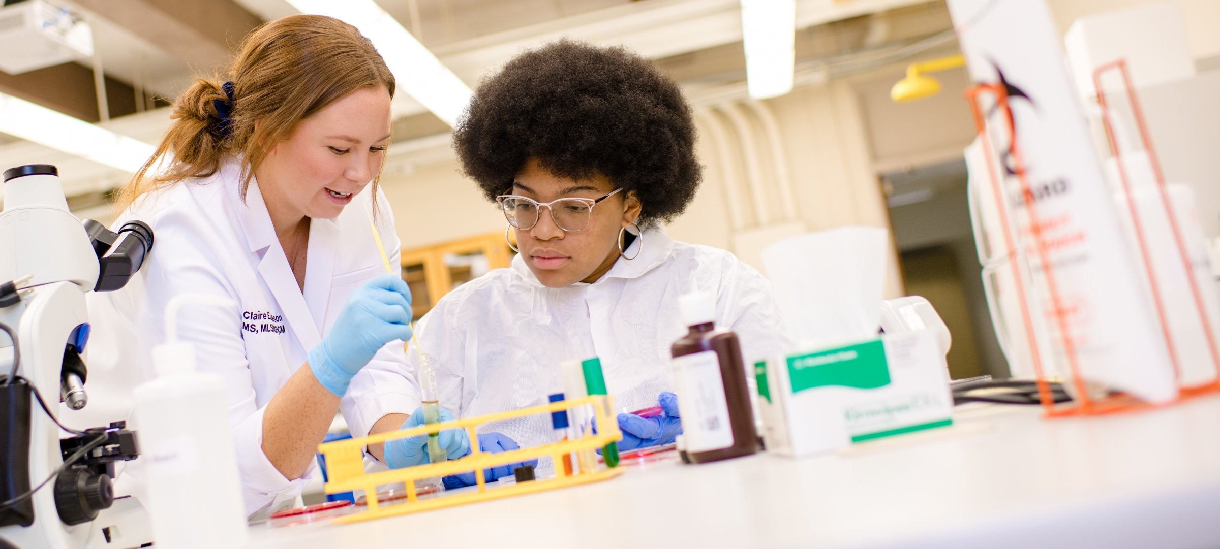 A white woman faculty member talks to a black woman student who is working with test tubes and vials which are on a table next to a microscope.