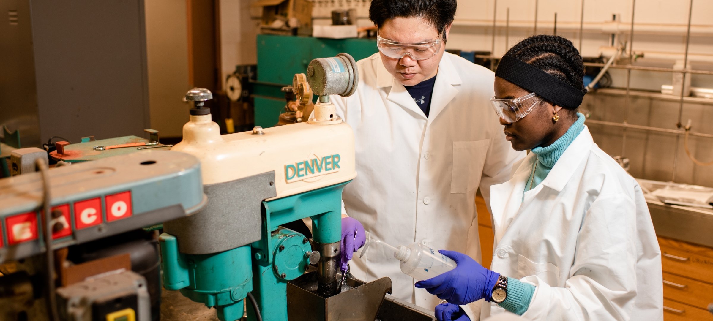A black woman student with braided hair holds a fluid filled bottle next to a machine while an asian faculty mentor observes. Both are wearing protective eyewear.