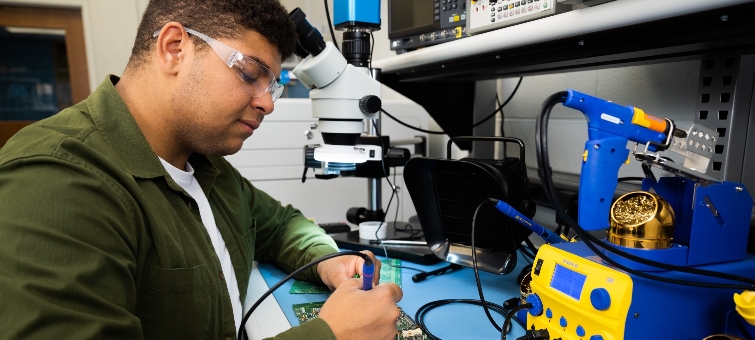 A student of color uses a soldering tool on a circuit board while wearing safety glasses.