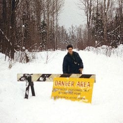 Man standing behind a baricade and a sign reading "Danger Area Authorized Personnel Only Beyond This Point" with additional words obscured by snow.