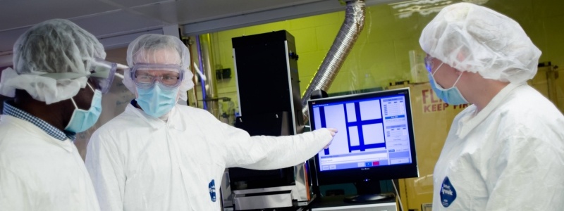 Three researchers in full protective gear in a lab with one pointing to a monitor