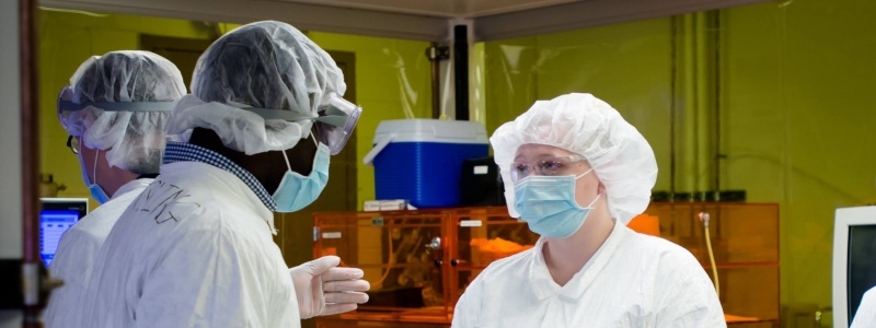 Two researchers discussing in lab in full protective gear with another researcher working in the background