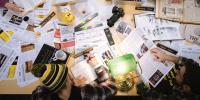 Papers, camera, water bottles clutter a table while students work around it.
