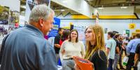 Gray-haired man in blue polo shirt speaks to female student wearing eyeglasses and smiling while holding an orange folder with resumes and employer information in a gymnasium with other students in business attire in background.