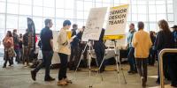 Students, faculty, staff, and community members walk around the atrium in the Van Pelt Opie Library near a sign that reads â€œSenior Design Teamsâ€ with a map of the room.