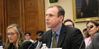 Stephen Techmann talks into a microphone while seated in a congressional meeting room, flanked on each side by other university experts in biotechnology.