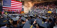 Looking toward the giant American flag behind the stage with many graduate in caps and gowns seated in front and people in the stands at commencement.