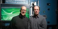 Raymond Shaw and Will Cantrell stand facing the camera in front of a green monitor screen mounted to the blue metal cloud chamber behind them.