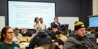 Three students share their SMART goals on a projection screen behind them as other students in the class look on.