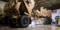 Two large tan tactical vehicles stand parked in a large warehouse structure, with the cab of the front vehicle folded up to show internal electrical components.