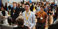 College students wearing yellow lanyards stand in lines to speak with company recruiters inside a gymnasium.