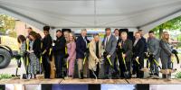 Campus leaders on a platform stage stand with black and gold shovels turning dirt during a groundbreaking ceremony.