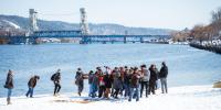 Students carry a concrete canoe from the snowy banks of the Keweenaw Waterway with the Portage Lift Bridge in the background.