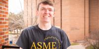Jacob Bruesch smiles in a black ASME T-shirt on an open balcony opposite the R. L. Smith Building.