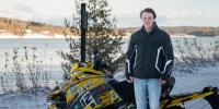 Jared Ott stands in front of a yellow MTU snowmobile on a snowy day near Princeâ€™s Point wearing a black jacket and blue jeans.