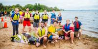 Ten Michigan Tech students pose on a beach with their cardboard boat, as other teams stage their boats in the background and race organizers survey the scene. There are sailboats moored in water in the background, along with a large orange buoy.
