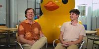 Professor Chuck Wallace and his student sit in a lounge with a large yellow rubber ducky in the background.