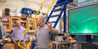 A man explains to a group of scientists the functions and capabilities of the machinery behind him, which includes a blue metal ladder and a large computer monitor displaying bright green waves of water particles.