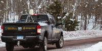 A black truck with the words Great Lakes Research Center and the Michigan Tech logo on the back drives away down a snowy road with an isotopic analyzer mounted in the truck bed.