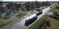 Ten trucks lay new pavement on a road through flat farmland in an aerial shot.