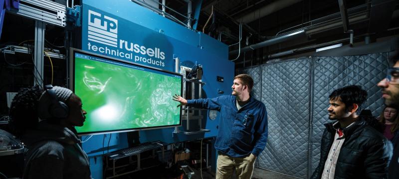 Students gathered around a professor pointing at a large screen with the russells technical products logo above it.